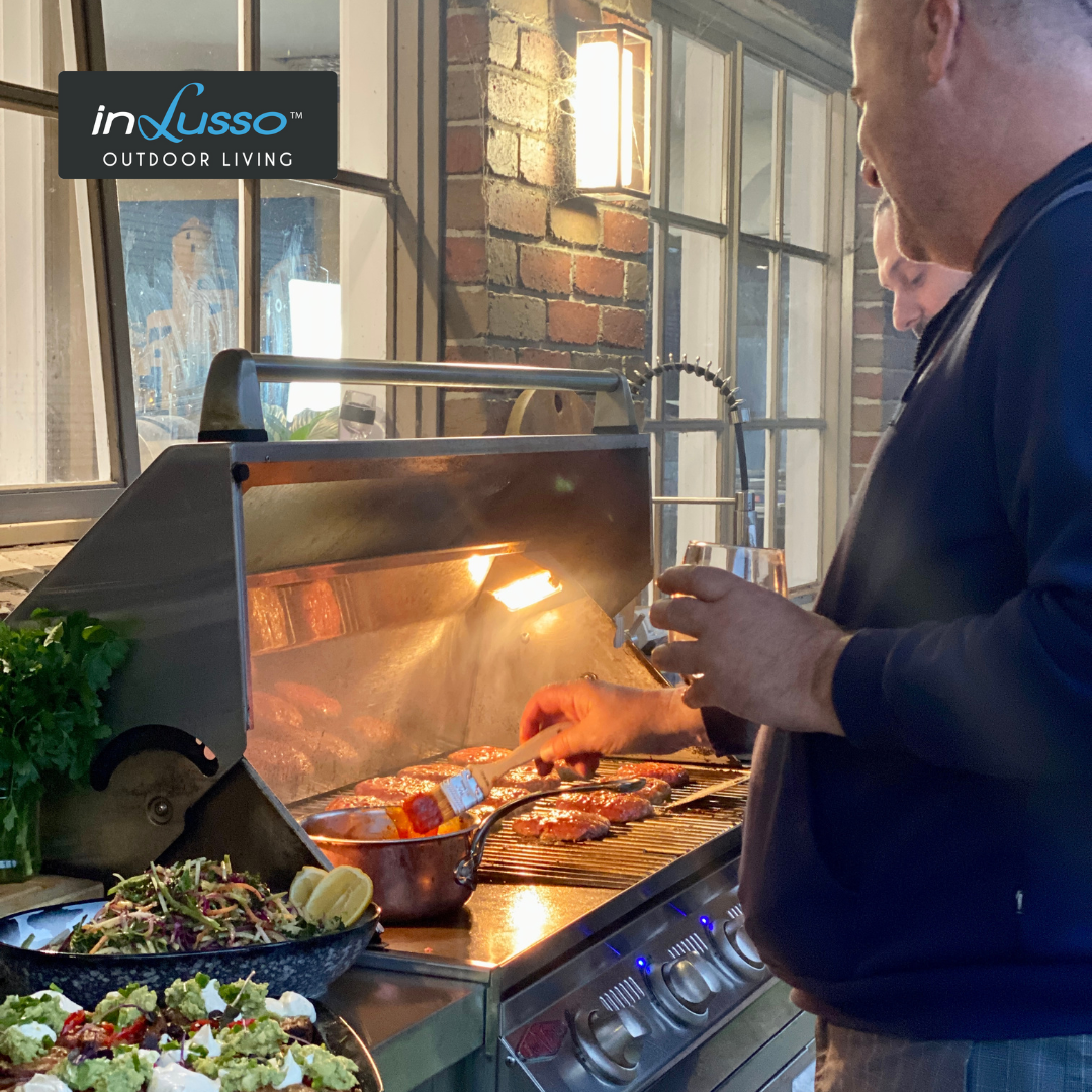 An outdoor BBQ kitchen with two men barbecuing burgers with glaze and salads in an alfresco area