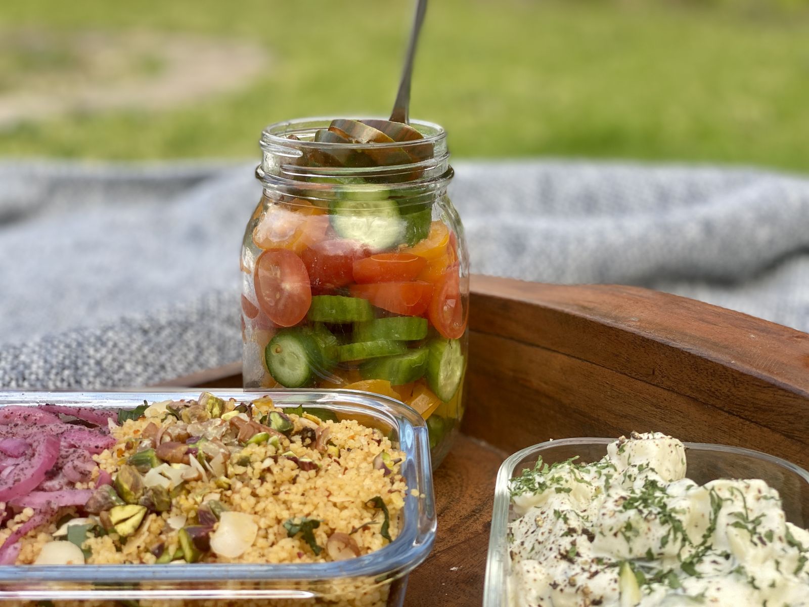 A jar with layers of mini heirloom tomatoes and baby cucumber slices on a picnic blanket with couscous and potato salad