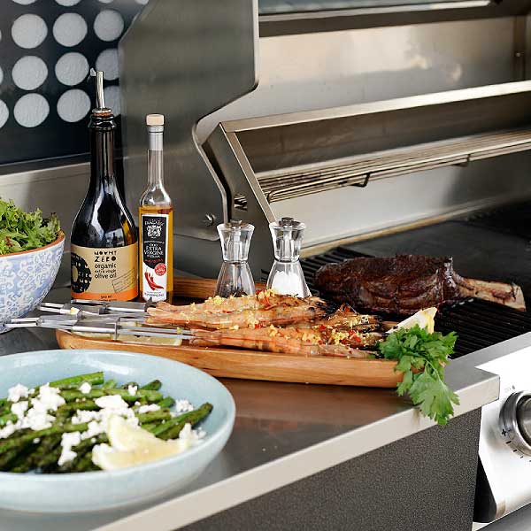 An outdoor BBQ kitchen with stainless steel bench, stainless steel hood, asparagus, prawns and steak.