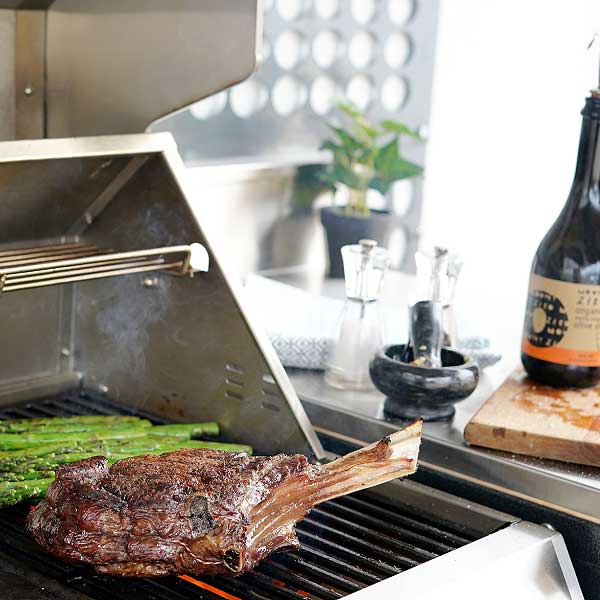 Outdoor BBQ kitchen with cast iron plates and hood, cooking a tomahawk steak with a styled kitchen bench top
